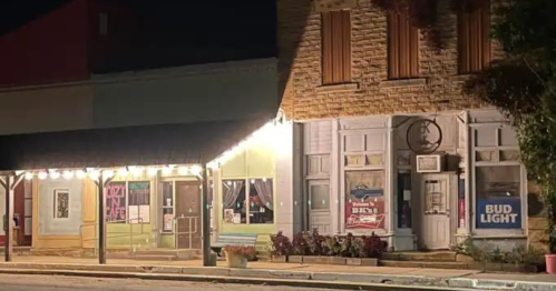 Nighttime view of a small town street with illuminated storefronts, including a bar and a shop displaying Bud Light.