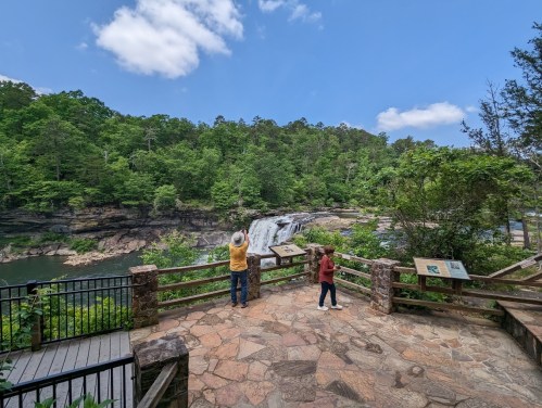 A scenic view of a waterfall surrounded by lush greenery, with two people observing from a wooden overlook.