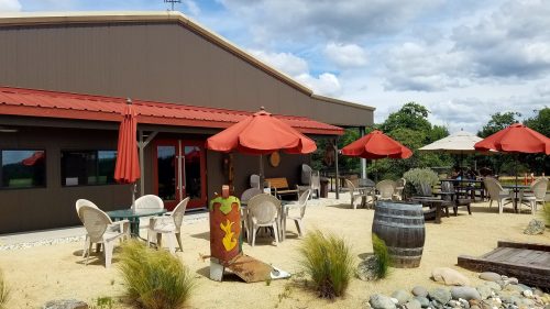 Outdoor seating area with umbrellas, a rustic building, and a barrel, surrounded by greenery and a sandy path.