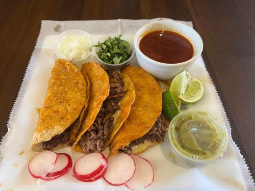 Three crispy tacos filled with beef, served with radishes, lime, cilantro, onions, and a side of red sauce.