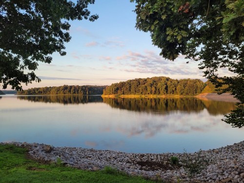 A serene lake at sunrise, surrounded by trees, with calm water reflecting the sky and shoreline.