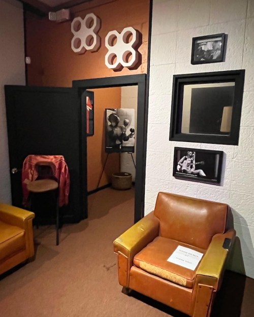 A cozy room with orange walls, two vintage chairs, and framed photos, leading to a darker hallway.