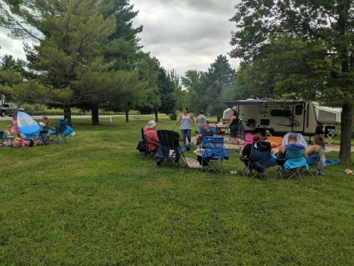 A group of people gathered around a campsite with chairs, an RV, and trees in a grassy area on a cloudy day.