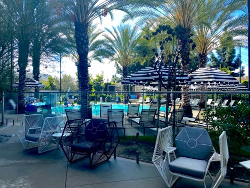 A poolside area with palm trees, striped umbrellas, and modern lounge chairs in a sunny setting.
