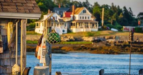 photo of a dock along the coastline in Maine during sunset, with a boathouse in the foreground and the water and houses along the shoreline in the background at sunset