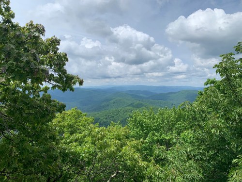 A panoramic view of lush green mountains under a cloudy sky, framed by trees in the foreground.