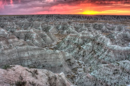 Sunset over rugged, layered badlands with dramatic rock formations and a colorful sky in the background.