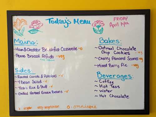 Whiteboard menu for April 14th featuring mains, sides, bakes, and beverages, with vegetarian options indicated.