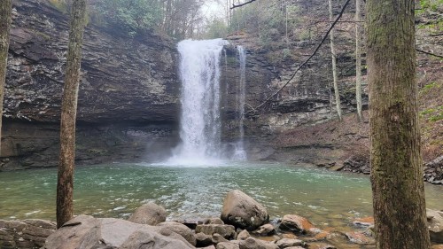A serene waterfall cascades into a tranquil pool, surrounded by rocky terrain and lush green trees.