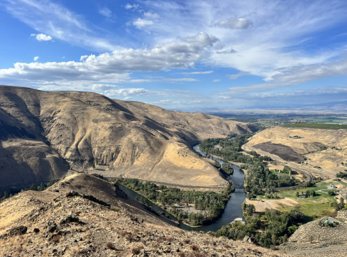 A scenic view of a winding river surrounded by dry hills and green patches under a blue sky with scattered clouds.