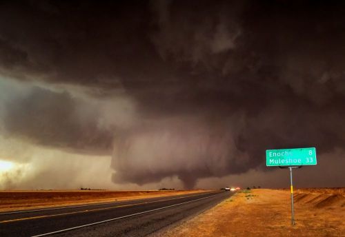 Dark storm clouds loom over a highway sign indicating distances to Enochs and Muleshoe, with a desolate landscape.