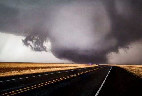 A dark, swirling storm cloud looms over a deserted road in a flat landscape, hinting at severe weather conditions.