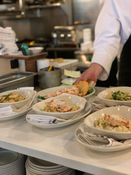 A chef serves pasta dishes with seafood in a restaurant kitchen, surrounded by plates and utensils.