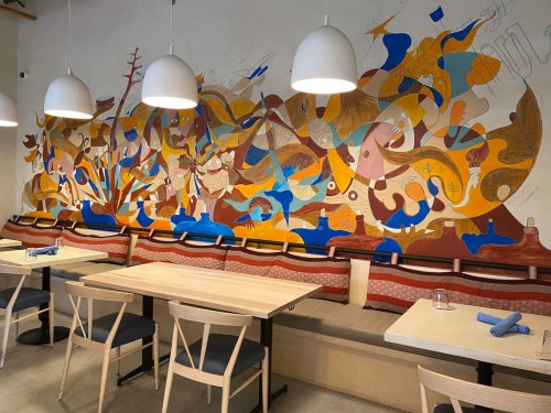 A colorful mural adorns the wall of a modern restaurant, with wooden tables and chairs arranged for dining.