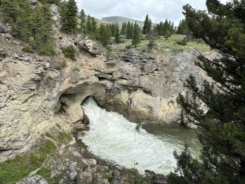 A waterfall cascades through a rocky cave, surrounded by lush greenery and trees in a mountainous landscape.