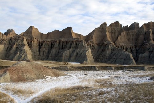 A rugged landscape featuring layered rock formations and a cloudy sky, with patches of snow on the ground.
