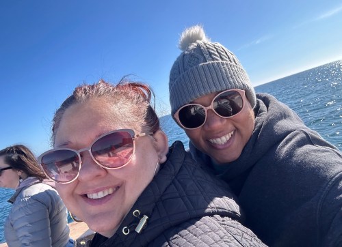 Two women smiling and posing for a selfie by the water, wearing sunglasses and enjoying a sunny day.