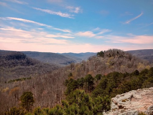 A scenic view of rolling hills and valleys under a blue sky with wispy clouds, showcasing a mix of trees and rocky terrain.