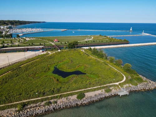 Aerial view of a coastal park with green fields, a pond, and a marina by the blue water under a clear sky.