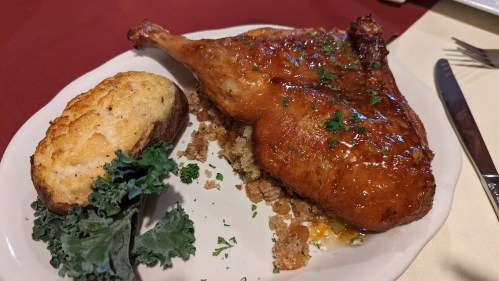 A plate featuring a crispy chicken leg with glaze, served on a bed of stuffing, alongside a slice of bread and kale.