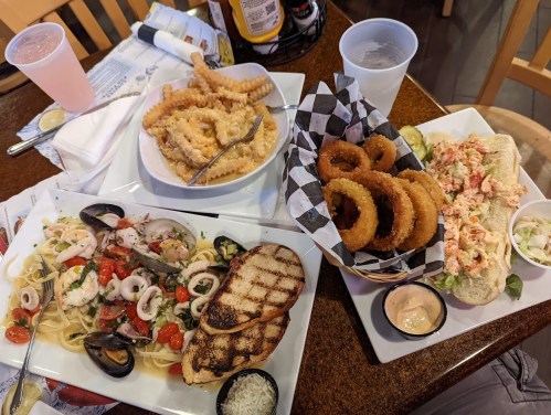 A table with two plates of seafood pasta, a lobster roll, onion rings, and crinkle-cut fries, with drinks in the background.