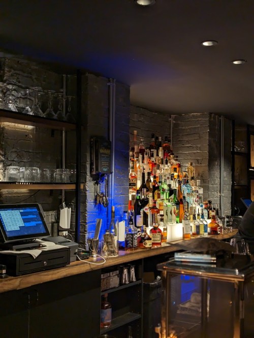 A dimly lit bar with a variety of colorful liquor bottles stacked against a brick wall and a cash register on the counter.