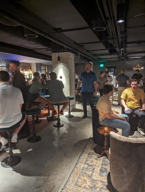 A lively bar scene with people sitting and standing, chatting, and enjoying drinks in a modern, dimly lit space.