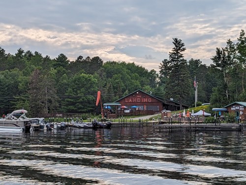 A lakeside view of a building labeled "The Landing," surrounded by trees and boats on calm water under a cloudy sky.