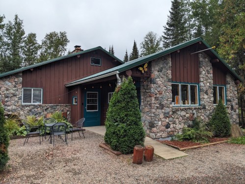 A rustic stone and wood cabin surrounded by trees, featuring a gravel path and outdoor seating.