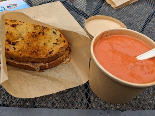 A grilled cheese sandwich on brown paper next to a cup of tomato soup with a plastic spoon.