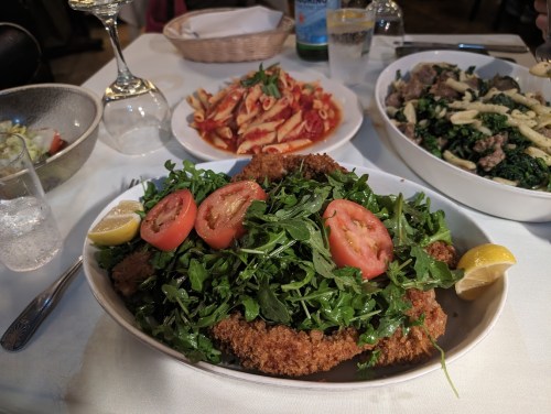 A plate of crispy fried chicken topped with arugula and tomatoes, surrounded by pasta and a vegetable dish.
