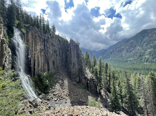 A scenic view of a waterfall cascading down rocky cliffs, surrounded by lush green trees and mountains under a cloudy sky.