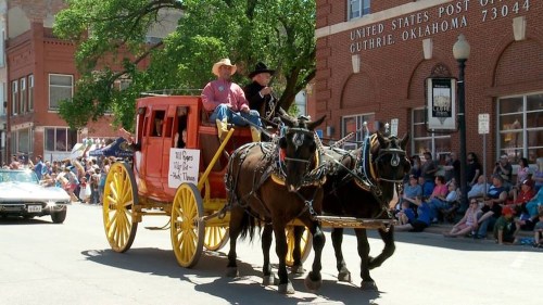A horse-drawn wagon with two riders parades down a street, surrounded by spectators and historic buildings.