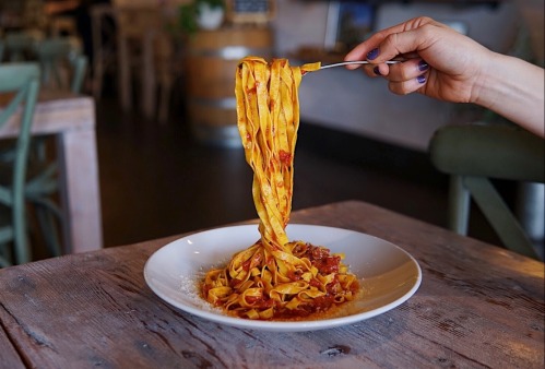 A hand holds a fork with long strands of pasta above a plate of spaghetti on a rustic wooden table.