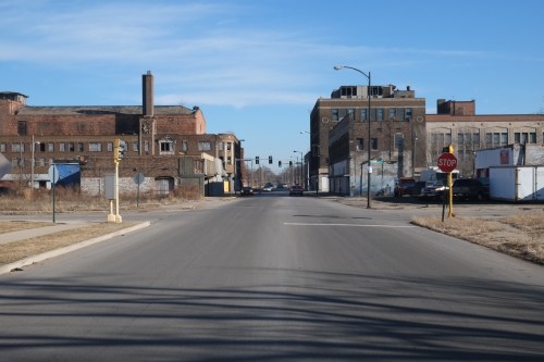 A wide, empty street lined with old industrial buildings under a clear blue sky. Traffic lights and a stop sign visible.