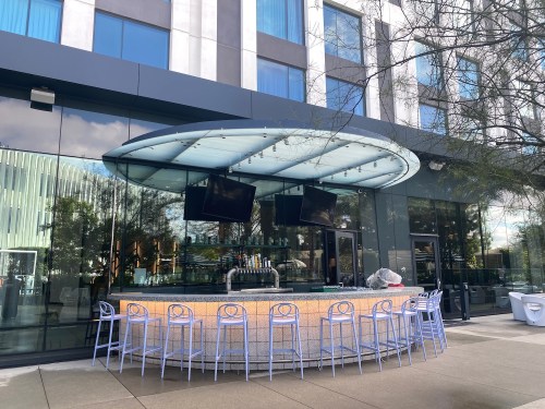 Outdoor bar with a curved roof, surrounded by light blue stools, next to a modern building with large glass windows.