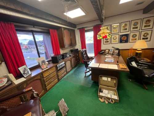 A vintage office with a wooden desk, red curtains, green carpet, and framed awards on the wall.