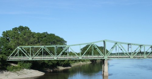 A green truss bridge spans a calm river, surrounded by lush trees under a clear blue sky.