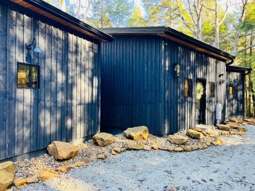 Modern black wooden cabin surrounded by trees, featuring large windows and a stone pathway.