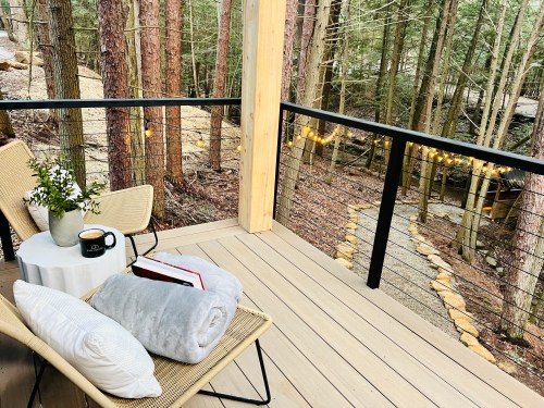 Cozy outdoor seating area with a blanket, coffee, and a book, surrounded by trees and soft string lights.