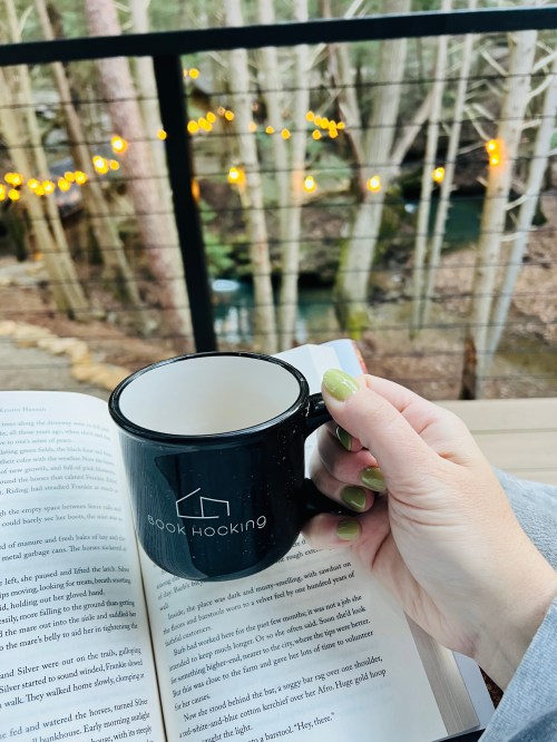 A hand holding a black mug over an open book, with a scenic view of trees and string lights in the background.