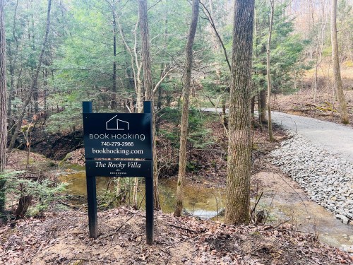 Sign for "The Rocky Villa" by Book Hocking, surrounded by trees and a gravel path along a creek.