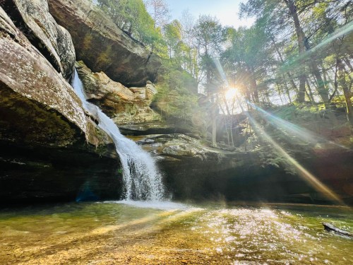 A serene waterfall cascades into a clear pool, surrounded by lush greenery and sunlight filtering through the trees.
