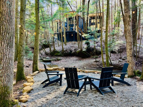 A serene forest scene with four black chairs arranged in a circle on gravel, surrounded by trees and a cabin in the background.