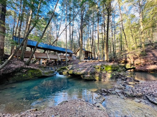 A serene forest scene featuring a wooden structure by a clear stream, surrounded by tall trees and rocky terrain.