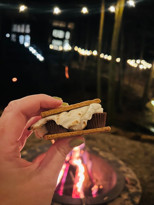 A hand holds a s'more with a toasted marshmallow and chocolate, with a glowing campfire and string lights in the background.