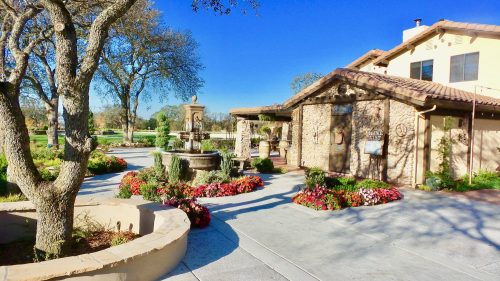 A charming stone building with a fountain, surrounded by colorful flowers and trees under a clear blue sky.