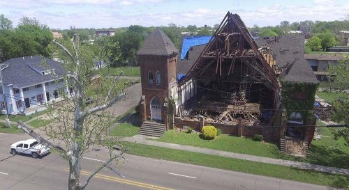 A damaged church with a collapsed roof, surrounded by greenery and a street, with a police car nearby.