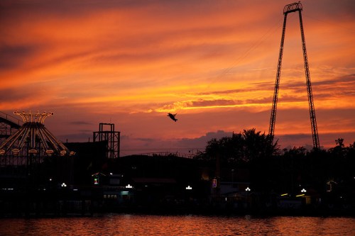 Silhouette of a person on a swing ride against a vibrant sunset sky, with amusement park structures in the background.