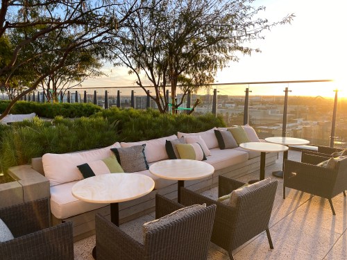 Rooftop lounge area with plush seating, small tables, and greenery, overlooking a sunset cityscape.